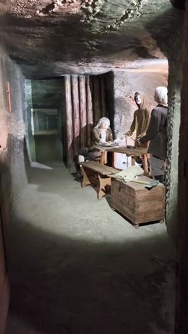 The video shows a downward angle inside a dark stone structure, possibly a basement or tunnel. On the left is a rough stone wall. In the foreground, a weathered wooden door. In the center, a steep wooden staircase leads down into the darkness, with a glimpse of a lit lower level at the bottom. Above the stairs are mannequins dressed in historical garb, operating a large wooden contraption.