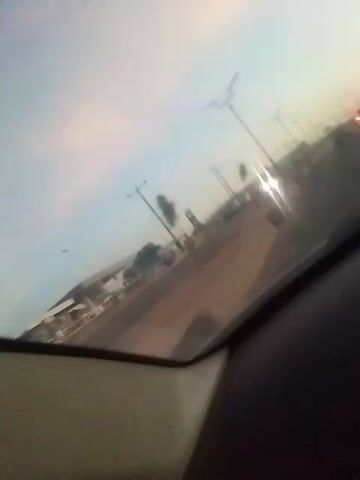 The video shows a view through a car windshield, with the dashboard visible at the bottom. A road stretches out, lined with streetlights and utility poles. A gas station is visible to the left. Several cars are present on the road. The sky is overcast with a hint of pink near the horizon, suggesting dawn or dusk.