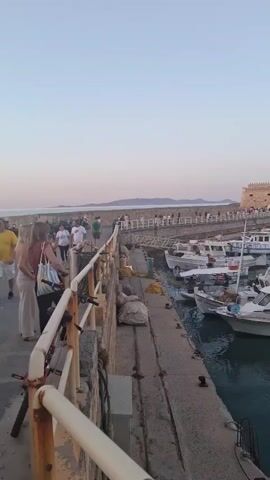 The video presents a view of a harbor, likely in the early evening or late afternoon, as the light is soft. A stone pier is visible in the foreground, with a white railing lining its edge. Several people are walking along the pier. The harbor is filled with a variety of boats, from small fishing boats to larger yachts. In the background, a stone fortress stands, with people visible along its ramparts. Further out in the harbor, there are more boats and what appears to be a larger ferry or cruise ship docked. The sky is a pale blue, and the water is calm, reflecting the sky and the boats. The overall scene is peaceful and scenic.