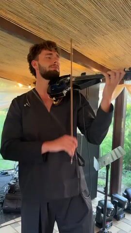 A young man with brown curly hair and a beard, wearing a black outfit with an open collar, plays an electric violin. He is standing indoors under a thatched roof with bamboo-like covering. There are string lights overhead. In the background, there is a stage with musical equipment including speakers, cables, and light fixtures. Another person, partially visible, is to the left, holding a microphone. The man then switches to playing a black and orange accordion, also called a Bugari accordion. The accordion has a decorative orange and black pattern on its front. He plays the accordion for a few seconds. The scene appears to be an outdoor performance setting.