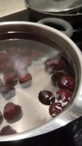 A close-up shot shows dark red cherries being boiled in a silver pot filled with water. Bubbles rise to the surface around the cherries, indicating that the water is hot. The pot is on a black stovetop, and a lid is visible in the background. The lighting is dim, suggesting the scene may have been filmed indoors in the evening or in a poorly lit kitchen. The focus is on the cherries as they cook, with some of them appearing slightly translucent.
