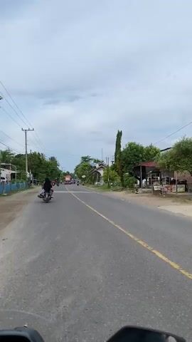 The video shows a view from a vehicle traveling down a road. On the left side of the road, there is a sidewalk, a blue fence, houses, trees, and utility poles. On the right side of the road, there are buildings, trees, and more houses. Several motorcycles are visible on the road. In the distance, there is a red truck, and other vehicles. The sky is cloudy. The road has yellow and white lines marking the lanes. A person on a motorcycle wearing a black headscarf is in the foreground on the left. A person on a motorcycle wearing a white shirt and helmet is in the foreground on the right. The overall impression is of a rural or suburban area with moderate traffic.