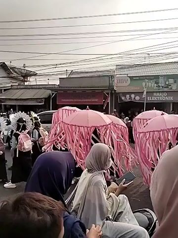 A group of people are gathered outdoors on a street. Several individuals are holding large, pink umbrella-like structures decorated with pink fringe, resembling jellyfish. Some people are dressed in costumes, including head coverings and what appear to be face masks. Spectators are visible in the foreground, some seated and looking towards the parade. The background shows buildings and overhead power lines, indicating an urban or semi-urban environment. The overall scene suggests a festive event or parade.