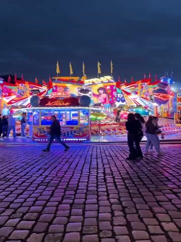 This video captures an amusement park ride at dusk. The ride is brightly lit with colorful flashing lights, predominantly red, yellow, and blue. It appears to be a spinning or swinging ride with multiple arms and platforms. Several people are visible on the cobblestone ground in front of the ride, some walking, others standing and observing. The sky is dark, indicating it is nighttime or late evening. The ground is paved with cobblestones. The scene suggests a lively atmosphere at an entertainment venue.