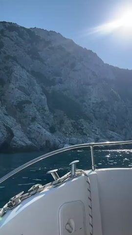 A first-person perspective video shows a boat moving along the coast of a rocky, mountainous island. The sun is shining brightly in a clear blue sky, casting a glare on the water and the side of the boat. The boat is white with silver railings and has ropes and cleats visible on the deck. The rocky cliffs are steep and covered in sparse vegetation, with some white patches suggesting rock slides or exposed stone. The water is a deep blue and appears choppy with sunlight reflecting off its surface. The boat appears to be moving at a moderate speed. There are no people visible in the video.