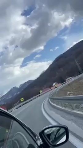 The video shows a car driving along a winding road in a mountainous area. The road is paved and has guardrails. There are trees on the mountainsides and power lines along the road. The sky is cloudy with patches of blue. Other cars can be seen on the road. The camera is positioned inside the car, looking out the driver's side window.