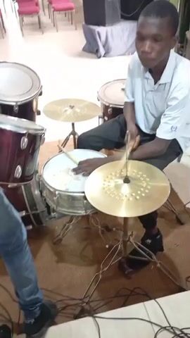 The video shows a young person playing a drum set. The person is seated and holding drumsticks. They are hitting a snare drum and a cymbal. Another person's arm and shoulder are visible on the left side of the frame. The drum set is on a rug. In the background, there are chairs and speakers. The lighting appears to be indoor lighting. The camera is positioned at a medium shot, capturing the drummer's upper body and the drum set. The video appears to be a short clip of a drumming performance or practice session.