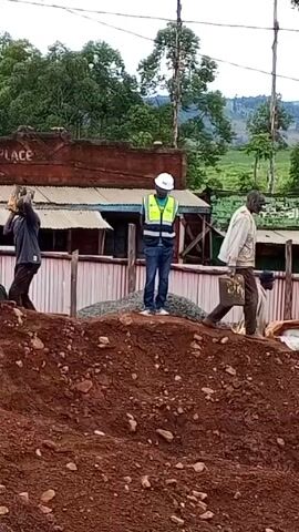 The video shows a construction site with several workers. They are building on a mound of reddish-brown dirt. One man stands atop a pile of gravel, wearing a hard hat and reflective vest, seemingly overseeing the work. Other workers are carrying buckets and bags, moving materials around the site. In the background, there is a building with the words "THE SALUTE PLACE" visible on its facade. The surrounding environment includes trees, power lines, and what appears to be a tea plantation on the distant hills. The sky is overcast.