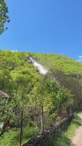 The video shows a waterfall cascading down a green, lush hillside. The waterfall is the primary focus, with water flowing over rocks and through trees. The sky is blue and mostly clear. In the foreground, there's a weathered stone wall with a chain-link fence on top, and a dirt path runs alongside it. Some of the waterfall's top reaches appear to have some sort of small structure.