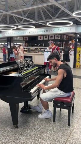 A young man with dark hair and a beard is playing a grand piano in a public indoor space. He is wearing a black sleeveless shirt and white shorts. He is seated on a red piano bench and his hands are on the keys. The piano is black and appears to be well-maintained. In the background, there are people walking and standing. There is also a bakery with a display of food items. The lighting in the space is bright, with circular fluorescent lights on the ceiling. The overall atmosphere appears to be busy and casual.