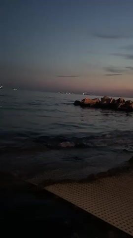 The video shows a view of a body of water, likely the sea, at dusk or dawn. The sky is a deep blue with hints of pink and purple near the horizon. The water is calm with gentle ripples. In the distance, several ships or boats with lights are visible. On the right side of the frame, a cluster of large rocks juts out into the water. In the foreground, a textured surface, possibly a deck or pier, is visible. The lighting suggests it is twilight. There are no people visible in the video.