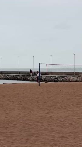 Two people are on a beach with a volleyball net. A person in a white top and blue jeans approaches the net, followed by a person in a grey top and red pants. The person in the white top and blue jeans appears to be a female. They both stop at the net, with the person in the grey top and red pants standing closer to the net. There are several people walking on a concrete wall in the background, which is next to the ocean. There are light poles on the concrete wall. The beach is sandy and appears to be light brown in color. The sky is overcast and grey.