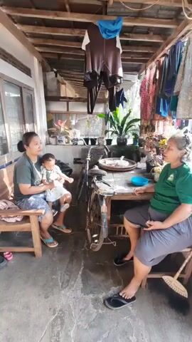 Two women and a baby are sitting in a covered outdoor area. The woman on the left is holding the baby. The woman on the right is sitting at a table. There is a bicycle behind the table. Clothes are hanging on a line above them. The ground is concrete.