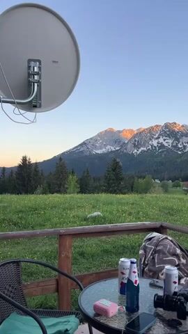 From a fixed camera on a cabin porch, a picturesque mountain vista unfolds under a clear sky. The mountain range in the distance is partially snow-capped, with golden sunlight illuminating its peaks. A lush green meadow stretches toward the mountains, dotted with clusters of wildflowers and trees. The porch itself is furnished with a glass-topped table surrounded by wicker chairs with green cushions. On the table, there are several beverage cans, a bottle, a digital camera, two smartphones, and a pink device, possibly a vape or a small charger. A folded blanket is draped over the back of one of the chairs. A satellite dish is mounted above the porch, partially visible in the top left corner of the frame. The scene overall conveys a sense of relaxation and appreciation of the natural beauty.