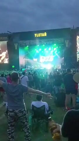 A wide shot shows a large crowd gathered in front of a stage at an outdoor concert. The stage is brightly lit with spotlights and features a large screen displaying a band's name. People are standing and sitting, some with their arms raised in excitement. The sky is overcast.