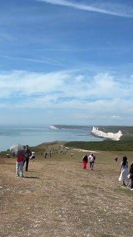 The video shows a scenic coastal landscape with white cliffs and the sea. A group of people are walking on a grassy hill. The sky is blue with some clouds. The people are dressed in casual clothing. Some are walking individually, while others are in small groups. One person in the foreground is holding an umbrella. The white cliffs are a prominent feature in the background, stretching along the coastline. The sea is calm and blue. The overall atmosphere appears to be one of leisure and tourism.