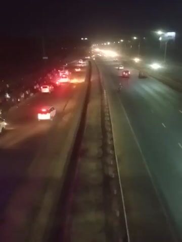 A nighttime scene shows a road with several vehicles stopped, some with their hazard lights on. One bus is overturned on its side, with people gathered around it. Another bus is parked nearby, illuminated by purple lights. The road appears wet, reflecting the lights from the vehicles and overhead streetlights. A line of vehicles is visible further down the road. The overall impression is of an accident scene with an overturned bus.