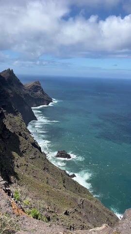 The video shows a view from a cliff overlooking the ocean on a partly cloudy day. The cliff is rocky and covered in some vegetation.  The ocean is a deep blue color, with white waves crashing against the rocky coastline.  The camera slowly pans across the scene to the right, showing more of the vast ocean expanse.