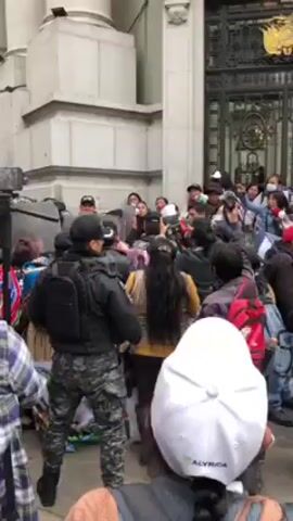 The video shows a dense crowd of people gathered in front of a large, light-colored building. The crowd is predominantly composed of civilians, some wearing traditional clothing, interspersed with uniformed police officers.  The police are equipped with shields, forming a line to contain or block the crowd. Some members of the crowd are pushing against the police line, and one person can be seen using a megaphone. A person on the crowd can be seen filming with a phone on a tripod. The environment appears to be an urban setting, with the building suggesting an important government or public institution. The overall atmosphere suggests a protest or demonstration taking place.