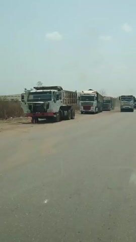 The video shows several trucks parked on the side of a paved road under a mostly clear sky. The closest truck, a white dump truck, is parked on the shoulder of the road, and it appears to be somewhat old and worn. Behind it, there are three more trucks; two of them have trailers. All the trucks seem to be facing the same direction. The road appears to be dusty and has some cracks. Beyond the trucks, the landscape is relatively flat with some sparse vegetation visible. The video seems to be taken from inside a moving vehicle.