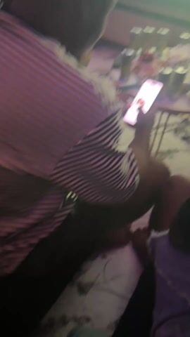 The video shows a group of people indoors, likely at a gathering. A person wearing a striped shirt is looking at a phone. A small round table is cluttered with multiple cans, bottles, and food containers. A large screen in the background displays a soccer game. Another person is visible on the right side of the frame wearing dark clothing and light-colored denim shorts. The floor is marble-patterned. The lighting is dim.