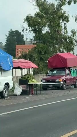 The video shows a roadside stall with a red pickup truck. The truck has a large red canopy on its roof. Several green and white t-shirts, presumably soccer jerseys, are hanging on display in front of the truck, along with several Mexican flags. In the background, there are trees, buildings, and a hillside with more structures. The overall scene appears to be a commercial setup along a road, likely selling merchandise related to a sporting event or national pride.