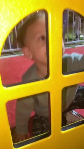 The video captures a young child interacting with a bright yellow play structure, possibly a part of a miniature house or ride. Initially, the child's lower legs, clad in dark patterned pants, and a light-colored top are visible through the arched and square cutouts of the yellow panel. The ground beneath appears to be a red carpet or mat.

As the camera pans up, the child's hands, with red nail polish on one finger, are seen gripping the top edge of the yellow structure. The child, a toddler with light brown, curly hair, then looks down intently, their face partially obscured but showing a focused expression. A dark smudge is visible on their upper lip.

The background is dark and blurred, revealing indistinct shapes of people and subtle lights, suggesting the scene takes place at night or in the evening. A metal railing is also discernible behind the child. The overall setting appears to be a lively, public event or amusement area, possibly an outdoor fair or indoor play space. There is no visible text in the video.