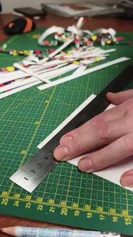 The video shows a close-up of a person's hands using a craft knife and a metal ruler to cut thin strips of paper on a green cutting mat. The mat has a grid pattern with yellow markings. The strips of paper appear to be cut from a larger sheet that has a colorful, patterned edge. There are many cut strips piled up to the left of the cutting area. A pencil with a patterned design is visible at the bottom of the frame. In the background, out of focus, are some office supplies, including a computer mouse. The lighting suggests an indoor setting, possibly during daylight hours. The activity depicted is precise paper cutting, likely for a craft or art project. There is no visible text in the frame.