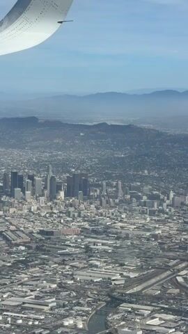 This is a vertical, aerial video shot from the window of an airplane. The airplane's wing is visible in the upper left quadrant of the frame. Below the wing, a vast urban landscape stretches out, characterized by dense buildings and a network of roads. In the middle ground, a cluster of skyscrapers forms a distinct downtown area. In the background, a hazy range of mountains can be seen under a light blue sky. The overall impression is of a sprawling city viewed from a high altitude.