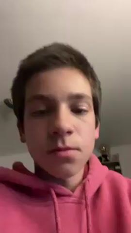 A person with brown hair and fair skin wearing a pink hoodie is looking at the camera. In the background, there is a partially visible ceiling fan and a shelf with various objects, including what appears to be a trophy. The person is positioned indoors.