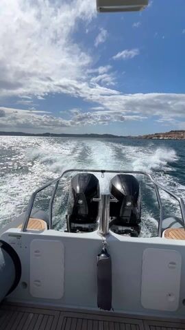 The video shows the view from the back of a white boat traveling across the water. Two black Mercury outboard motors are prominently displayed, creating a foamy wake behind the boat. The boat has a wooden deck and a silver railing. The sky is blue with scattered white clouds, and the sun is shining. In the distance, a coastline with buildings can be seen.