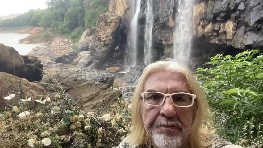 A man with long blond hair and a white beard is in the foreground of a video selfie. He wears glasses and a t-shirt with a pattern. Behind him is a waterfall with multiple streams cascading down a rocky cliff. The water flows into a river or lake surrounded by rocks and trees. The man gives a thumbs up gesture while wearing a red and black motorcycle glove.