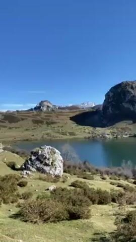 The video shows a scenic view of a lake surrounded by rugged mountains and lush greenery under a clear blue sky. Some of the mountain peaks in the background are covered in snow. The vegetation around the lake consists of low-lying shrubs and grassy areas. A large rock sits near the shoreline, adding to the natural beauty of the landscape. The camera pans slowly, offering a broader perspective of the lake and the surrounding mountainous terrain. There is the sound of people talking in the background.