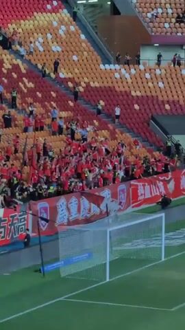 The video shows a soccer stadium with a section of fans wearing red shirts. The fans are behind a red banner that says "为重庆加油" which translates to "Cheer for Chongqing". The fans are waving flags and cheering. In the foreground is a soccer goal. The stadium seating is partially occupied and primarily colored in red and orange hues.