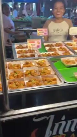 The video shows a food stall in an outdoor market at night. A woman stands behind a table covered with trays of fried food, possibly chicken, topped with green onions or herbs. Price signs are visible on some of the trays. Other stalls and people moving through the market are visible in the background. The sign on the front of the stall has writing in a non-English script. The stall is well-lit.