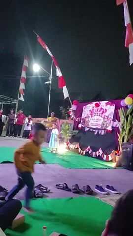 The video captures an outdoor nighttime event. In the foreground, a young child runs across the frame from right to left. The midground features a stage set up with a green carpet. A young girl in a light blue dress stands center stage, facing a microphone. Behind her, a man in a yellow shirt stands. To the left of the stage, several adults are gathered. A banner is displayed on the stage backdrop, decorated with red and white flags and balloons. The background shows trees and buildings under a dark sky. Multiple red and white Indonesian flags are prominently displayed, attached to tall poles on either side of the stage. In the lower part of the frame, several pairs of shoes are placed on the ground, along with a water bottle. The lighting appears to be a mix of stage lights and ambient illumination.
