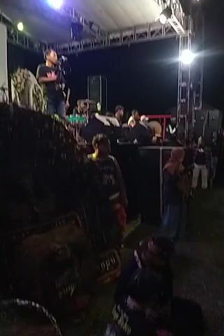 Okay, here's a description of the video:

The video appears to be shot at an outdoor event at night. It is very dark and the camera angle is tilted, making it difficult to see clearly.  At the start of the video, a stage with lighting and performers is visible in the distance. There are people standing in the immediate foreground, obscuring much of the view.  The camera moves slightly, revealing more of the crowd gathered in front of the stage. Some of the people in the crowd appear to be wearing masks. The event seems to be musical in nature due to the sound and the stage setup.