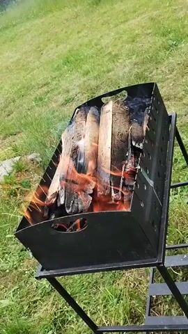 A fire is burning in a black metal grill. The grill is rectangular and has a stand with legs. It is placed outdoors on a grassy area. The fire consists of several pieces of wood, which are burning with orange flames and emitting smoke. The wood appears to be in various stages of combustion, with some pieces charred and others still brightly lit. The background is a blurry green expanse of grass, suggesting an outdoor setting.