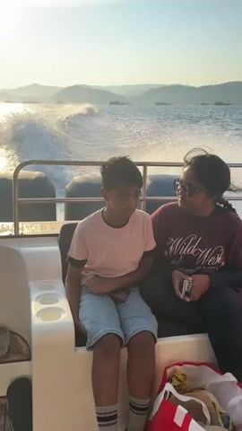 Two young people are sitting on the back of a boat as it speeds through the water. The sun is setting, casting a golden glow on the scene. The water is choppy, creating a white wake behind the boat. In the background, there are mountains and the silhouettes of ships. The boy is wearing a pink shirt and blue shorts, and the girl is wearing a maroon shirt.