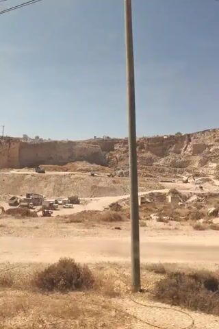 The video shows a quarry with heavy machinery, including excavators and trucks, engaged in excavation work. The landscape is arid and dusty, with piles of earth and rock. Overhead power lines are visible against a clear blue sky. In the background, there are buildings and structures, suggesting a nearby settlement or industrial area. The overall scene depicts an active mining or construction operation in a dry, rugged environment.