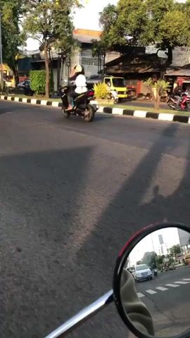 A motorbike rider, wearing a white helmet and a light-colored shirt, rides a motorbike on a paved road. The rider is in the middle of the frame, moving from left to right. Behind the rider, other motorbikes and a yellow car are visible. Trees and buildings line the side of the road. The sun appears to be setting, casting a warm light on the scene.