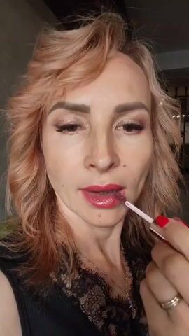 A woman with blonde hair and makeup is applying lipstick. She is wearing a black top with lace trim and a necklace. Her right hand is visible, holding the lipstick applicator. Her fingernails are painted red. The background is blurred and appears to be indoors.