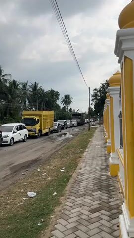 A stationary line of vehicles, including cars and trucks, is visible on a road with a sidewalk to the right. A yellow truck with a cargo bed is in the foreground, followed by several cars and other trucks. The road is paved, and there is a grassy area with some litter to the left of the road. The sidewalk is made of interlocking bricks. Behind the sidewalk, there is a decorative yellow and white wall with arched pillars. Buildings are visible in the background, including one with a sign that reads "SHOWROOM SUKSES LARIS MOTOR." Palm trees and other vegetation are also present. The sky is overcast with gray clouds.