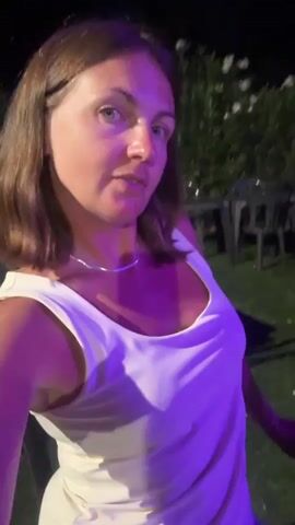 The video begins with a close-up of a woman with shoulder-length brown hair and blue eyes, wearing a white tank top and a silver necklace. She is lit by purple lights, creating a vibrant atmosphere. The background is dark and slightly blurred, with hints of outdoor seating and vegetation. The woman makes a few facial expressions, including a pout and a surprised look. In subsequent shots, she touches her hair and adjusts it. She appears to be at an outdoor event, possibly a concert or festival, as there are other people visible in the background, seated at tables and on benches, and a stage with lights can be seen. The woman waves to the camera in one shot. The overall scene suggests a lively and possibly festive outdoor gathering at night.