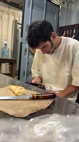 A young man with dark hair and a beard wears a white t-shirt and looks down at his smartphone. He is seated at a table with a slice of flatbread on brown paper and a knife with a decorative handle in front of him. The background shows a large gray shipping container, a wooden table with a water jug and metal pot, and a wall with kitchen utensils. The lighting is dim.