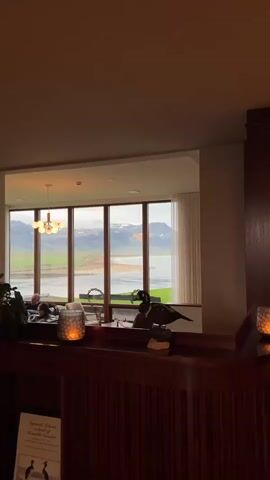 The video captures a tranquil interior scene offering a panoramic view of the dramatic Icelandic landscape outside. Inside, several people are seen relaxing in a cozy lounge or lobby area, seated on light-toned armchairs and engaging in conversation. The foreground features a dark wooden counter or bar, decorated with flickering candleholders that cast a warm glow. Prominently displayed on the counter is a dark, sleek bird statue, likely a guillemot or similar Icelandic sea bird. A modern, multi-bulb chandelier hangs from the ceiling, illuminating the interior.

Through the large windows, a breathtaking vista unfolds. Majestic, snow-capped mountains dominate the distant horizon under an overcast sky. In the middle ground, a calm body of water, likely a fjord or bay, stretches out with a dark, rugged shoreline. The weather appears cool and cloudy, but bright enough to clearly illuminate the stunning natural scenery.

The overall atmosphere is serene and comfortable, inviting relaxation. The time of day appears to be daytime, possibly late afternoon or early evening, with soft, diffused light. At the bottom left of the frame, partially visible text on a sign or brochure includes the words "Sjávargrettir," "Búðir," and "Ábyrgðarsjóður," confirming the location as Búðir, Iceland. The scene beautifully contrasts the warm, inviting interior with the raw, untamed beauty of the Icelandic wilderness.