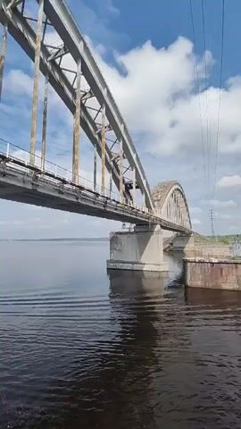 The camera is positioned on the water, possibly on a boat, looking at a large steel bridge. The bridge has a complex steel structure above the roadway, with rust visible on some of the metal parts. The roadway itself appears to be concrete and is supported by large concrete pillars in the water. The water is dark with slight ripples. The sky is blue with scattered white clouds. In the distance, on the far side of the bridge, there is a grassy shoreline and a tall utility pole.  The camera moves slowly forward, passing under the bridge.