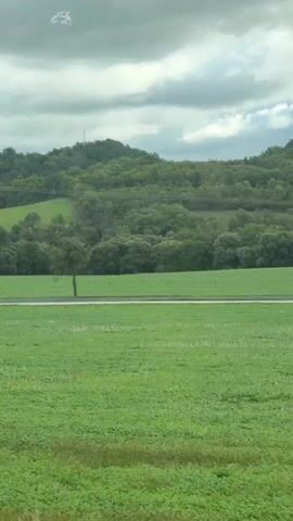 The video shows a view from a vehicle traveling through a rural landscape under a cloudy sky. The foreground consists of a green field. In the middle ground, there is a paved road with a single tree and a utility pole. Beyond the road, rolling green hills are covered with trees. The camera pans slightly to the right, showing more of the green fields and trees. The video is short and appears to be a time-lapse or a recording of a quick movement.