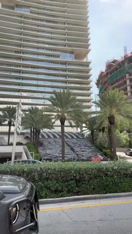 The video shows a tall, modern, light-colored apartment building with curved balconies. In front of the building, there is a cascading water feature with a striped pattern, surrounded by palm trees and lush green bushes. A blue shuttle bus is visible driving past the water feature and palm trees. A dark-colored SUV is parked in the foreground. To the right of the apartment building, a construction site with a crane and a partially built structure is visible. The sky is bright and appears to be daytime. A sign with the number "35" is visible on the left side of the frame.