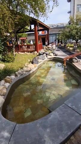 A stone-lined, shallow pool filled with murky, greenish water sits in the foreground of the video. The bottom of the pool is visible, covered with flat, irregular-shaped stones of varying shades of green and gray. A wooden walkway with a simple railing leads over the pool from the right side of the frame. The pool is surrounded by a lawn and large, dark gray and light-colored rocks. In the background, a red wooden pavilion with a dark roof stands to the left, featuring tables and chairs inside. To the right of the pavilion, a paved pathway leads towards buildings with traditional architecture, including paper lanterns hanging from the eaves. Trees with green foliage are present throughout the scene, casting shadows on the ground. The sky is visible in the upper portion of the frame, appearing partly cloudy with a bright, sunny atmosphere. No people are clearly visible in the immediate vicinity of the pool, though some figures can be faintly seen within the pavilion area. The overall impression is of an outdoor leisure or dining area with a water feature.